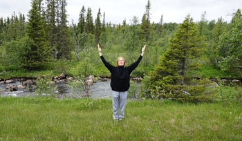 Person standing on grass with arms raised near a flowing river surrounded by green trees and forest under a cloudy sky.