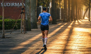 Person jogging on a sunlit paved path lined with trees, wearing a blue shirt, black shorts, white knee-high socks, and a black cap.