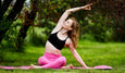 A person in athletic wear performs a yoga pose on a mat in a lush green outdoor setting, surrounded by trees.