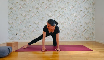 Person in black outfit doing a side stretch yoga pose on a red mat in a room with floral wallpaper and wooden floor.