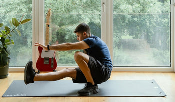Man in black shorts and navy shirt performing a one-leg squat on a fitness mat near a large window with a red guitar and a plant nearby.
