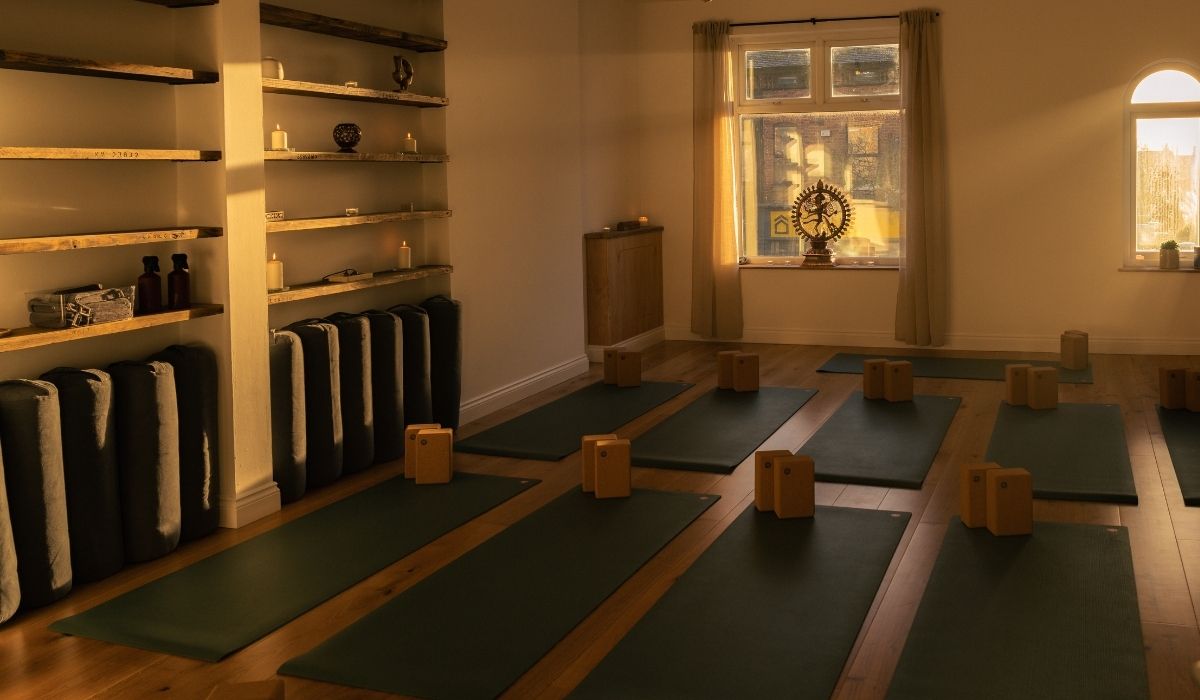 Serene yoga studio with wooden shelves, mats, blocks, and candles, bathed in warm afternoon light. Window features decorative elements.