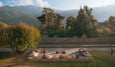 Outdoor seating area with cushions around a central fire pit, surrounded by trees and mountains in the background under a partly cloudy sky.