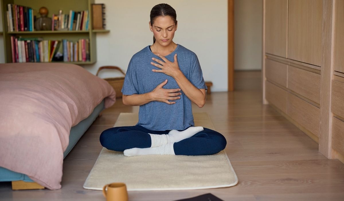 A person sits cross-legged on a mat, practicing mindfulness or meditation in a cozy, well-lit room with a bed and bookshelves.