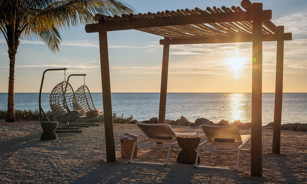A serene beach scene featuring hanging chairs under a wooden pergola at sunset, with the ocean reflecting warm hues in the background.