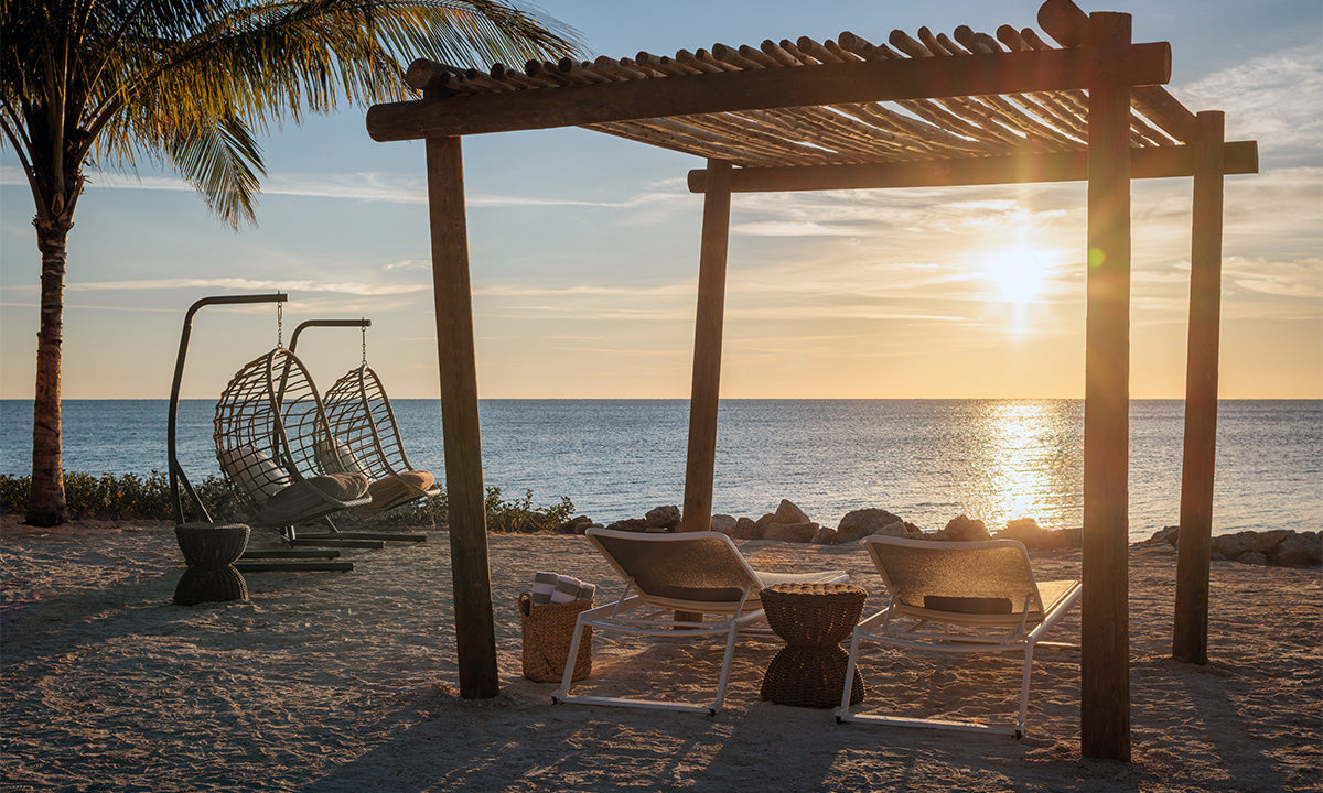 A serene beach scene featuring hanging chairs under a wooden pergola at sunset, with the ocean reflecting warm hues in the background.