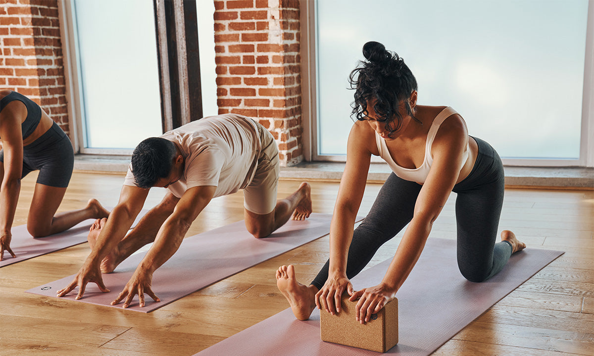 Three people practicing yoga poses on mats in a bright studio with large windows and exposed brick walls.
