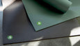 Two yoga mats in dark gray and green with textured surfaces, featuring a small green logo in the corner.