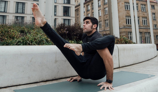 A person performs an advanced yoga pose on a mat outdoors, showcasing flexibility and balance against an urban backdrop.
