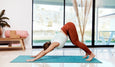 A woman in red leggings and a white top performs downward dog yoga pose on a mat, with plants and large windows in the background.