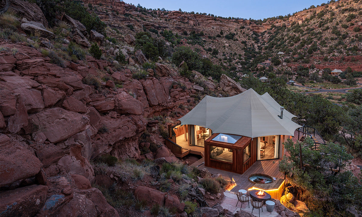 A modern tent-like structure nestled among red rocks, featuring a hot tub and outdoor seating, against a scenic canyon backdrop at dusk.