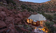 A modern tent-like structure nestled among red rocks, featuring a hot tub and outdoor seating, against a scenic canyon backdrop at dusk.