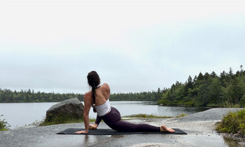 Person performing a yoga stretch on a mat on a rocky surface by a calm lake surrounded by forest under a cloudy sky.