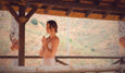 A woman stands in a serene outdoor setting under a thatched roof, practicing yoga with hands in a prayer position.