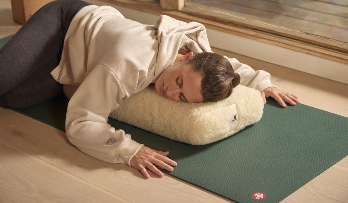 A person in a beige hoodie relaxes face down on a fluffy pillow, positioned on a green yoga mat in a sunlit room.