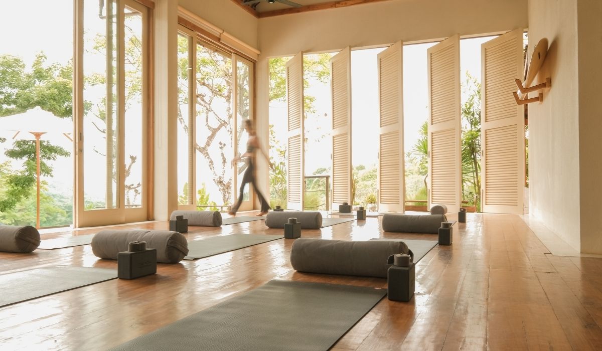 Bright yoga studio with large windows, yoga mats, bolsters, and a person walking, surrounded by lush greenery outside.