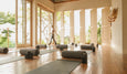 Bright yoga studio with large windows, yoga mats, bolsters, and a person walking, surrounded by lush greenery outside.