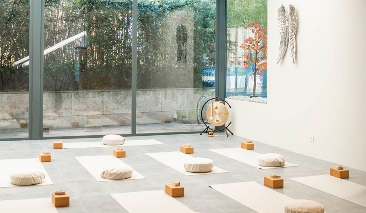 A serene yoga studio with meditation cushions, wooden blocks, and a gong, set against large glass windows and greenery outside.