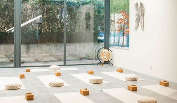 A serene yoga studio with meditation cushions, wooden blocks, and a gong, set against large glass windows and greenery outside.