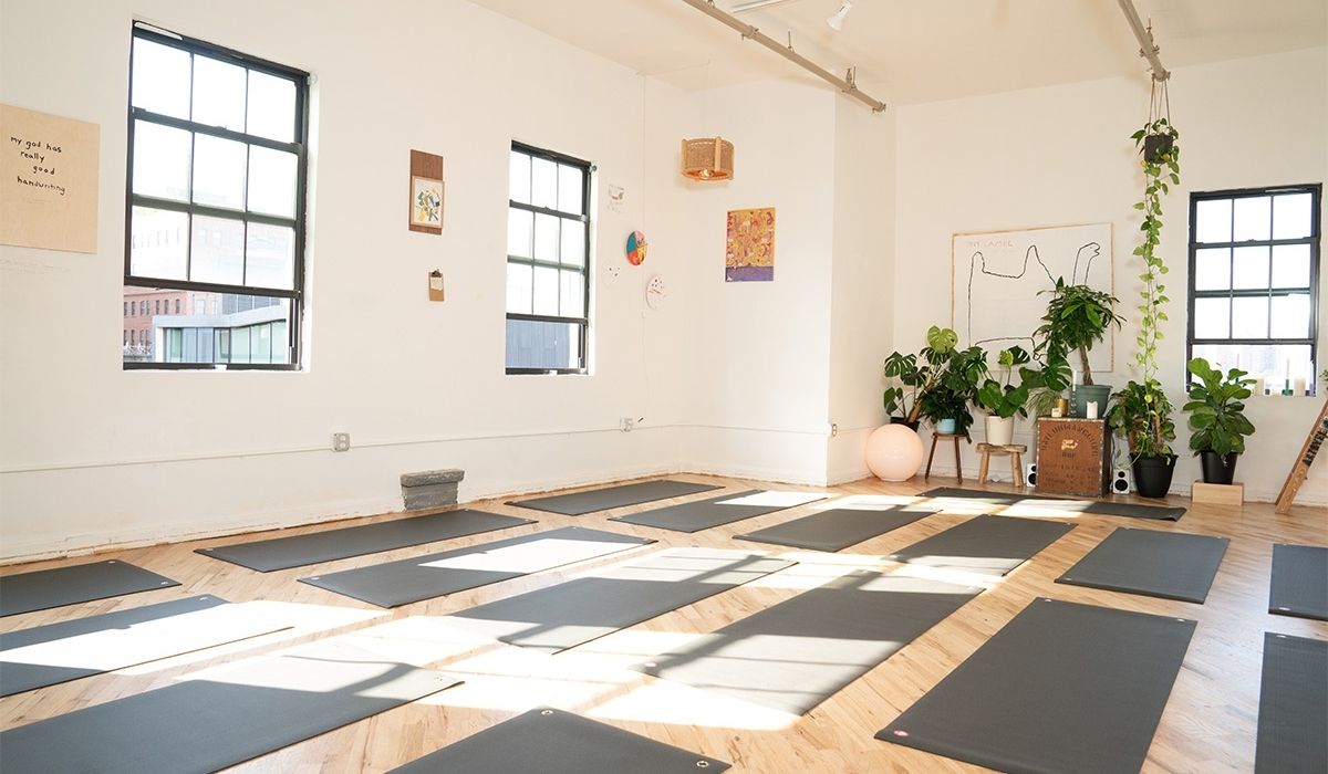 A bright yoga studio with wooden floors, lined with yoga mats, large windows, plants, and artwork on the walls.