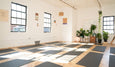 A bright yoga studio with wooden floors, lined with yoga mats, large windows, plants, and artwork on the walls.