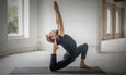 Man in black outfit performing a deep yoga stretch on a mat in a spacious, light-filled room with large windows.