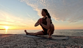 A person practicing yoga on a rock by the sea during sunset, with vibrant clouds and calm waters in the background.