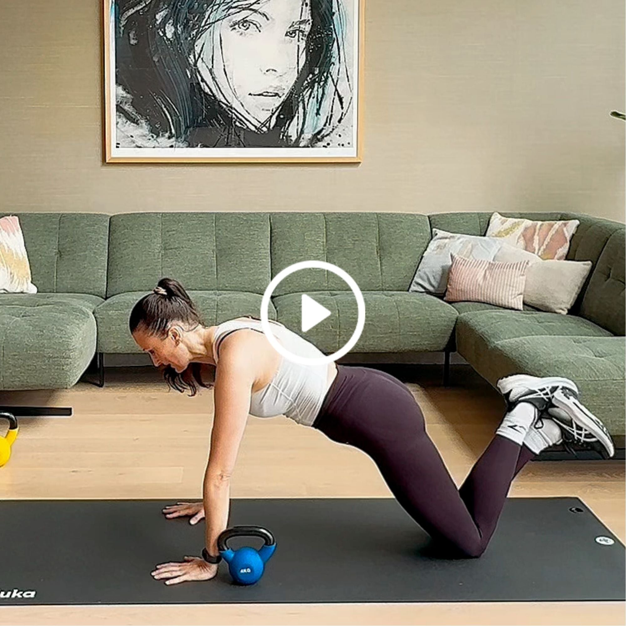 Woman in workout clothes doing knee push-ups on a black mat in a living room with a green sectional sofa and abstract face artwork.