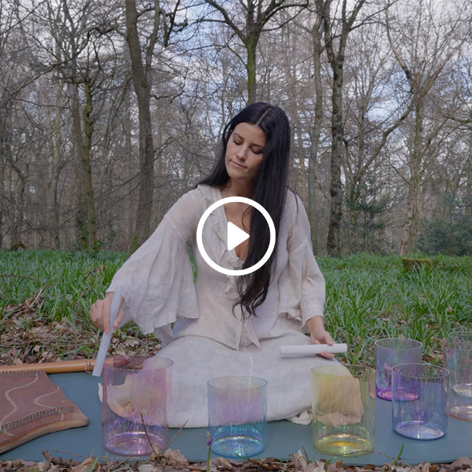 Woman in white dress playing crystal singing bowls in a forest clearing during daytime, surrounded by leafless trees and green grass.