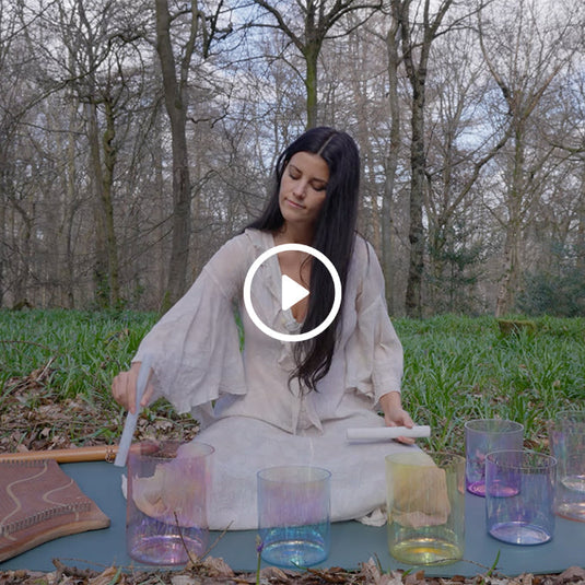 Woman in white dress playing crystal singing bowls in a forest clearing during daytime, surrounded by leafless trees and green grass.