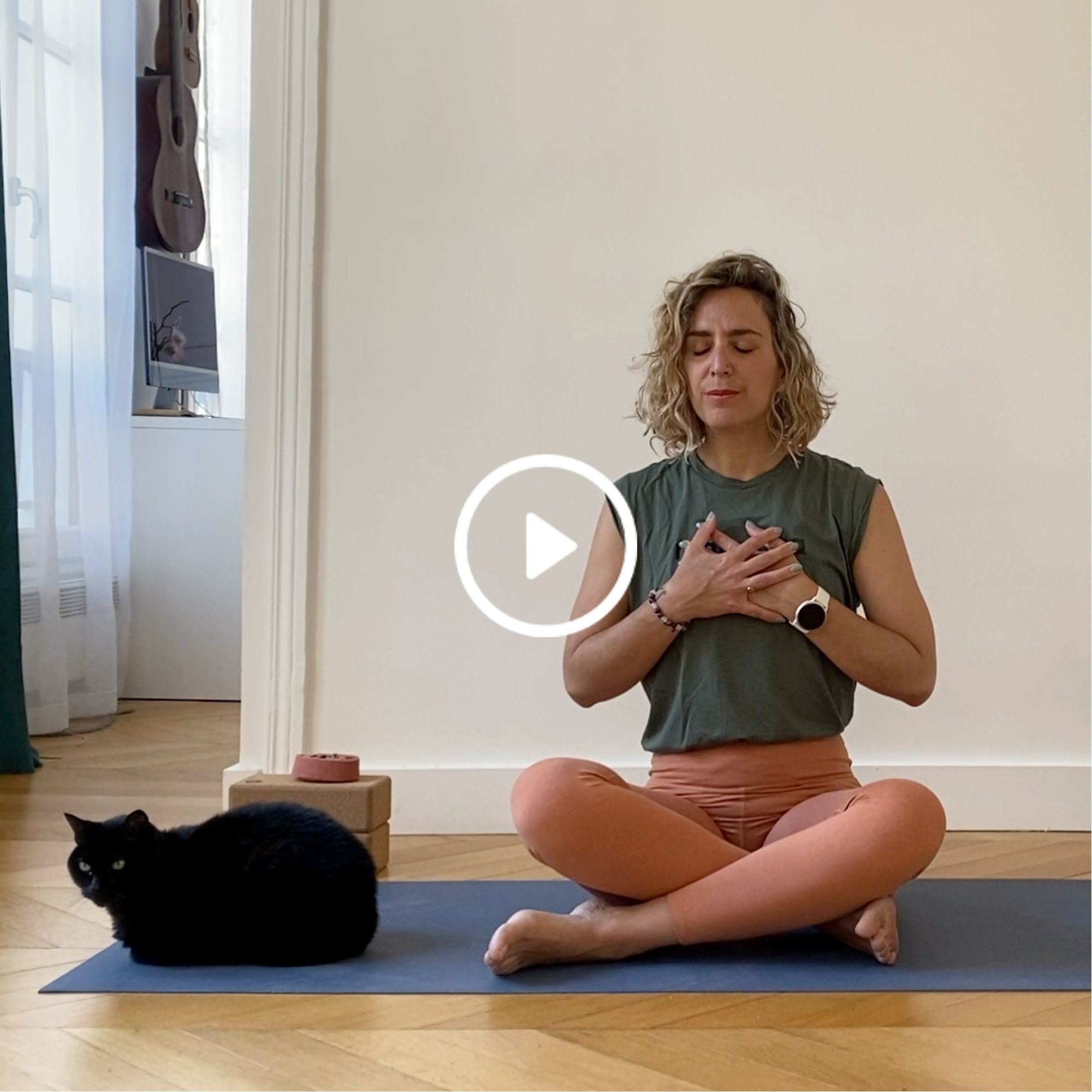 Woman in green top and orange pants meditating on a mat with a black cat sitting nearby in a bright room with wooden floor.