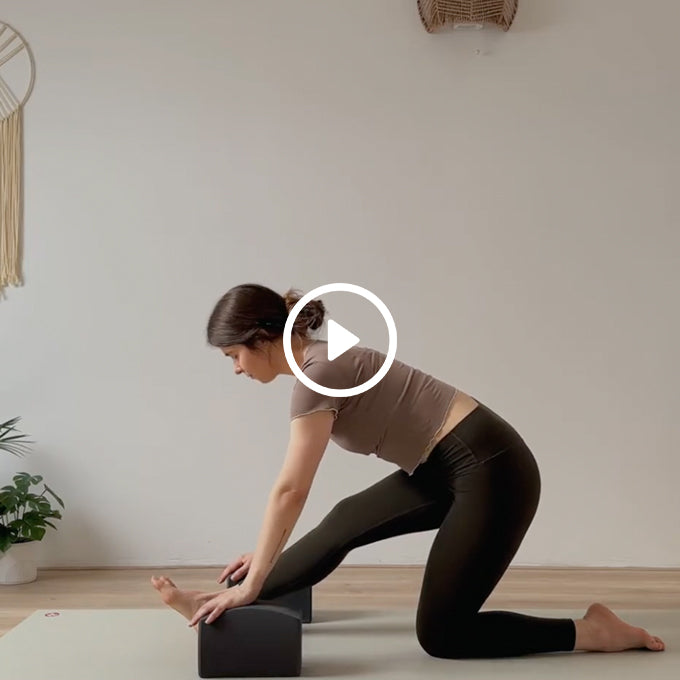 A woman in a brown top and black leggings stretching her hamstrings using yoga blocks on a mat in a minimalist room.