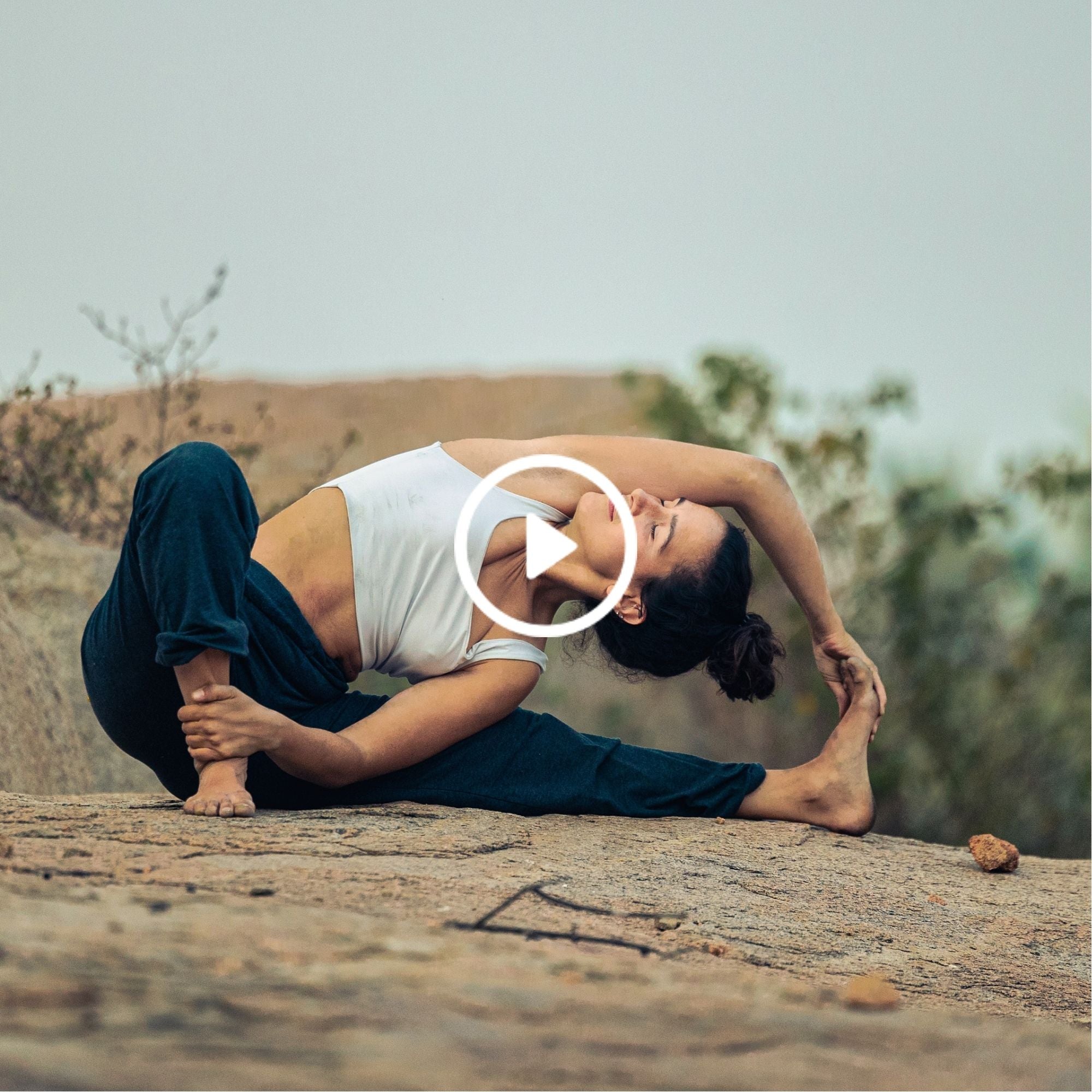 A woman in a white top and dark pants practices yoga stretching on a rocky surface outdoors with blurred greenery in the background.
