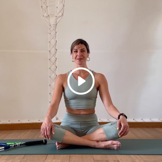 Woman in athletic wear sitting cross-legged on a yoga mat in a room with wooden floors and a tennis racket beside her.