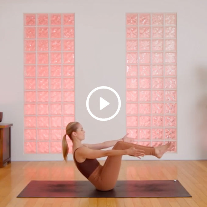 Woman in workout clothes performing a seated yoga pose on a mat in a room with pink-tinted glass block windows.