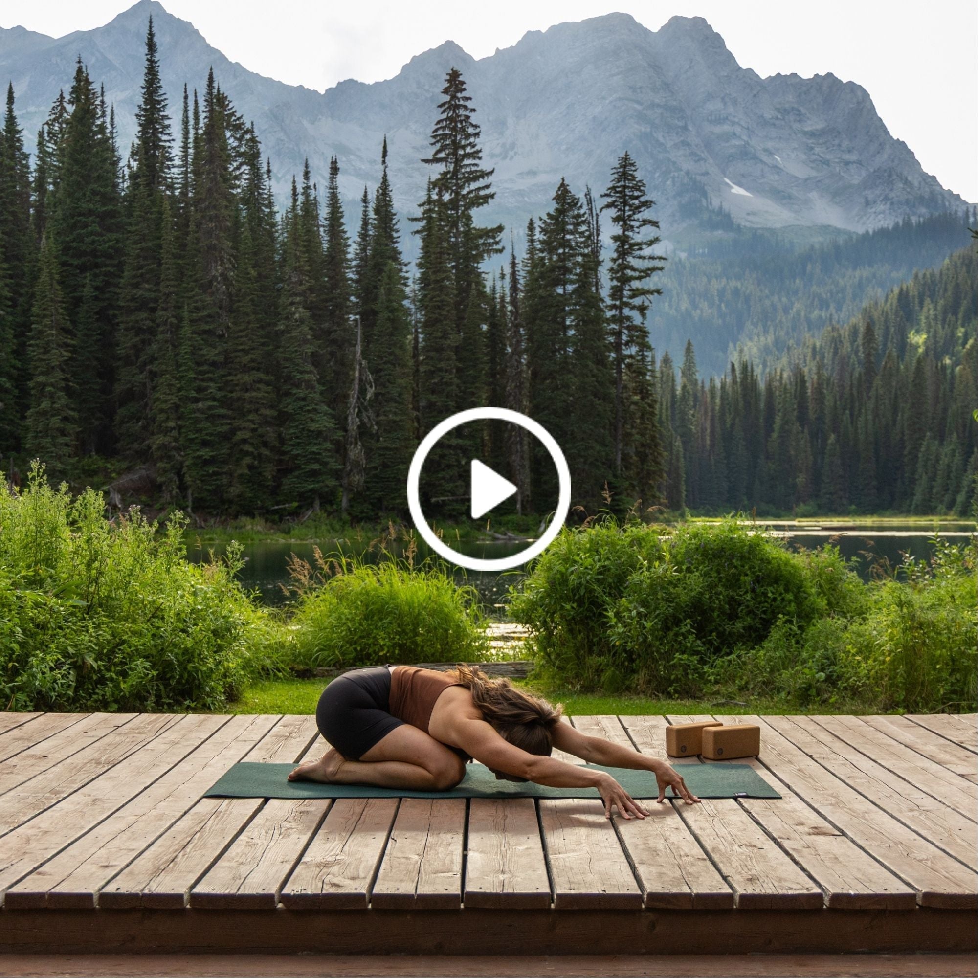 Person in child’s pose yoga on mat on wooden platform by lake, surrounded by lush greenery and tall pine trees with mountains in the background.