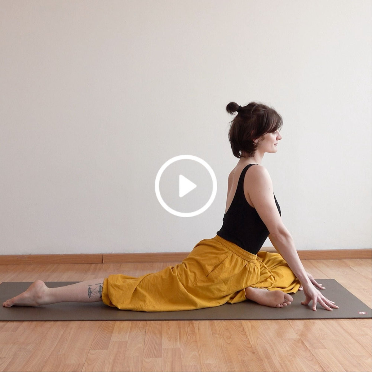 Person in a black top and yellow pants practicing a yoga split on a mat, with a play button overlay.