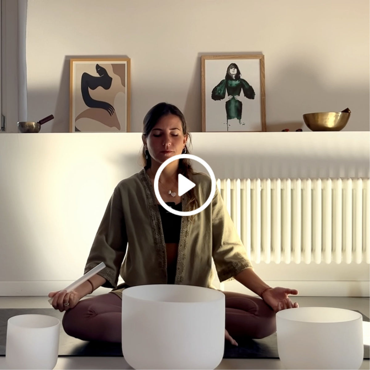 Woman meditating cross-legged behind crystal singing bowls, with a play button overlay and framed artwork in the background.
