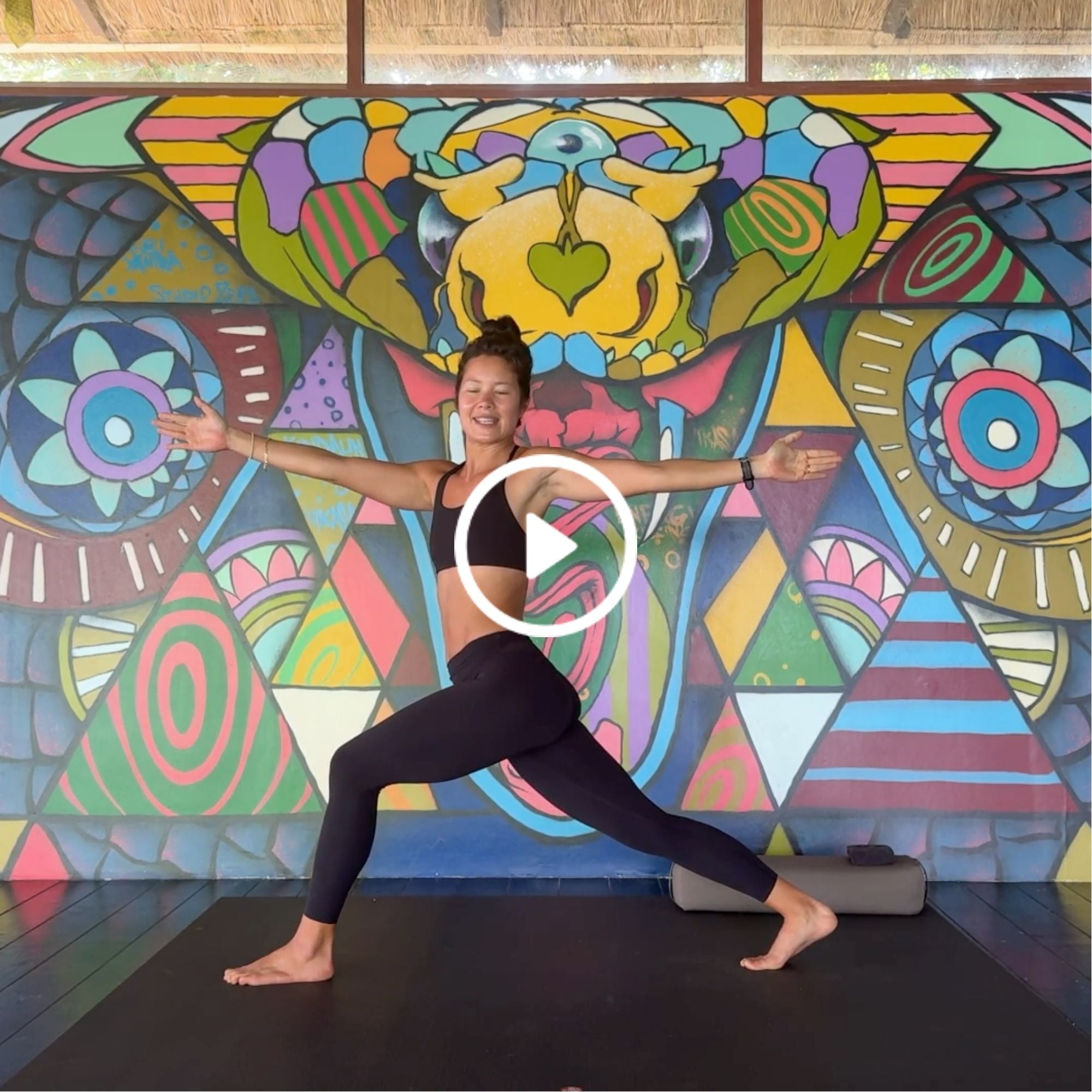 Woman in black athletic wear doing a yoga pose indoors with colorful, abstract mural featuring geometric shapes and vibrant patterns behind her.