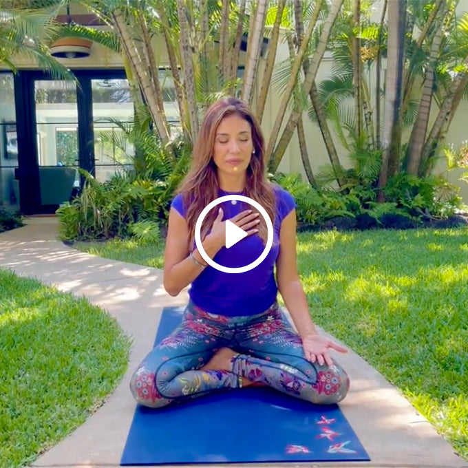 Woman meditating outdoors on a yoga mat with eyes closed, one hand on her chest and the other resting on her knee.