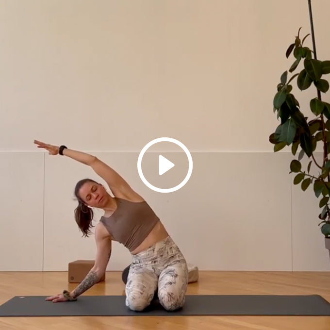 Woman in workout clothes performing a side stretch on a yoga mat in a minimalist room with a plant and yoga blocks nearby.
