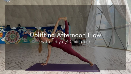 A woman in maroon workout clothes performs a side plank yoga pose on a purple mat in a spacious, bright studio with colorful wall art.