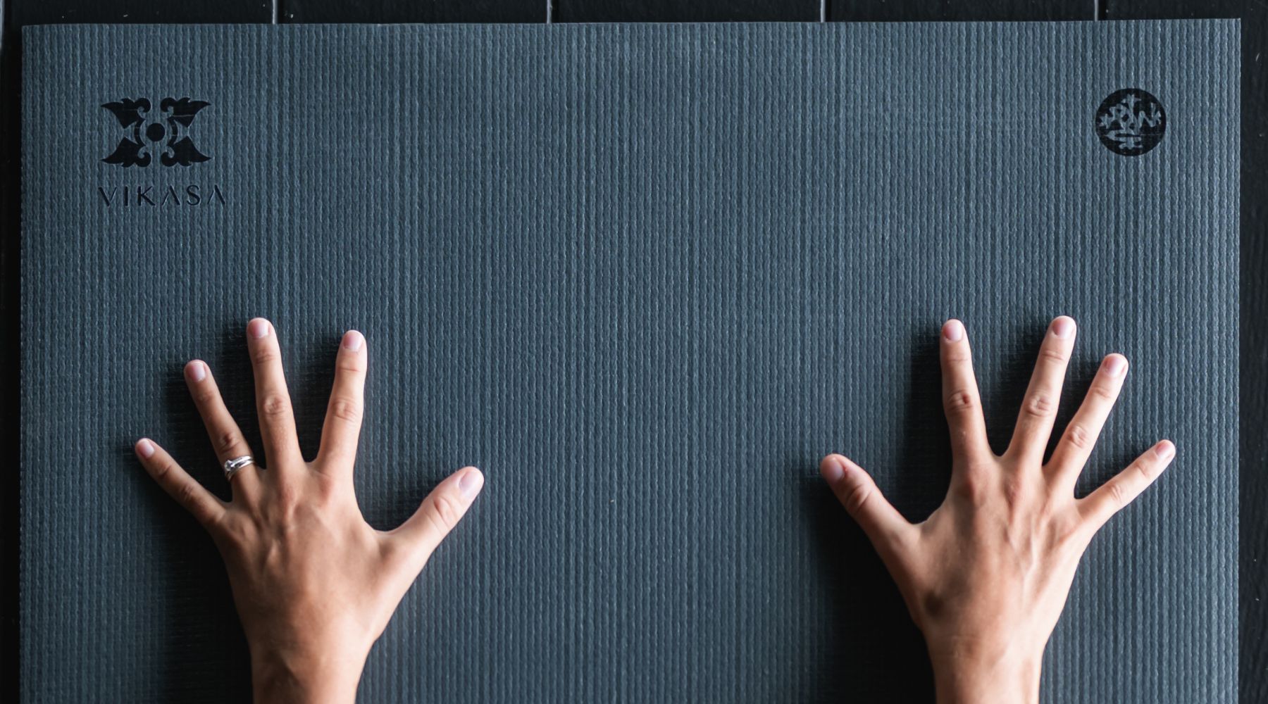 Two hands are placed on a textured Manduka PRO yoga mat in black thunder with the logo "Vikasa" visible in the top left corner.