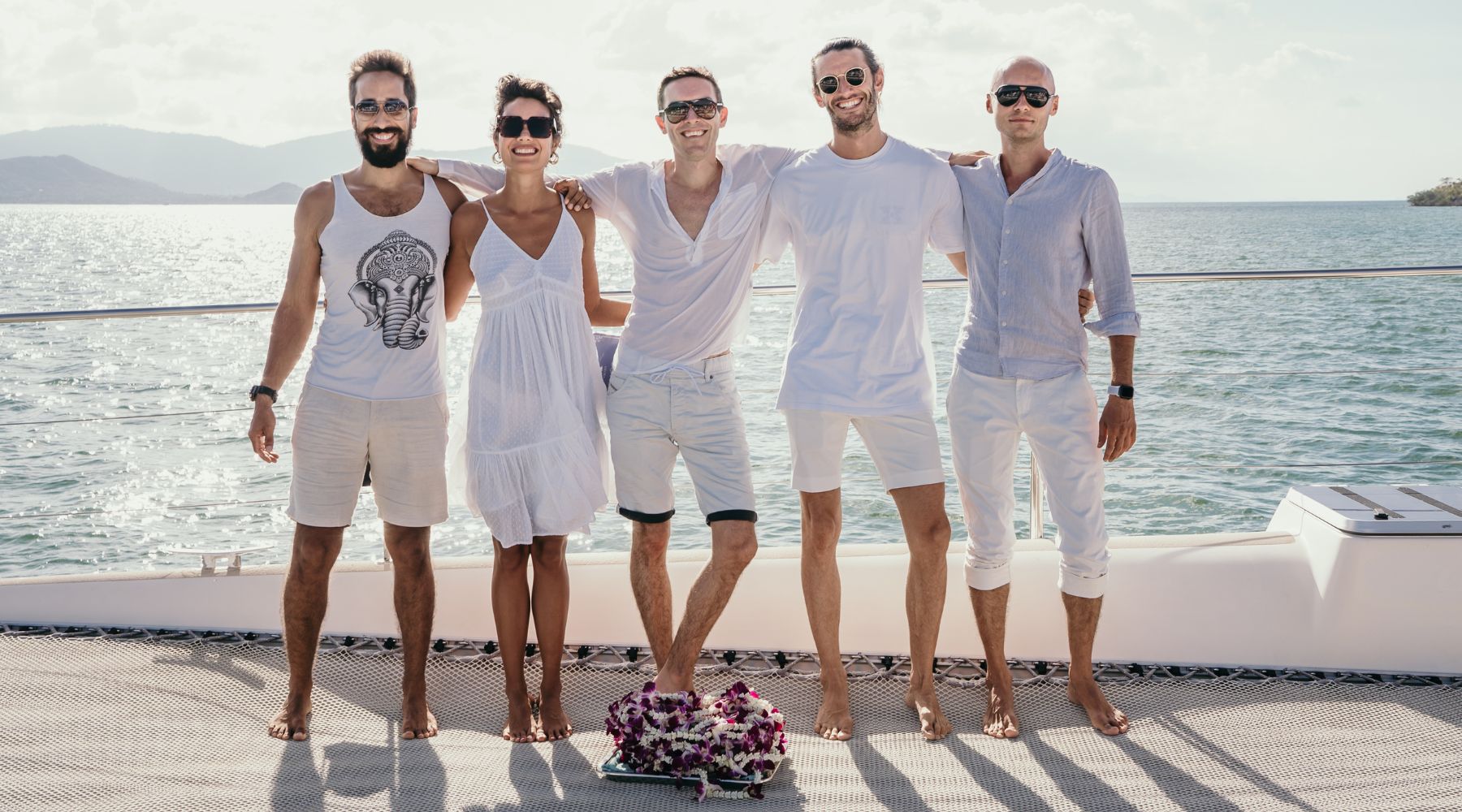 The Five Vikasa Yoga lead teachers pose together on a boat, wearing stylish summer outfits, with a scenic ocean backdrop and flowers at their feet.