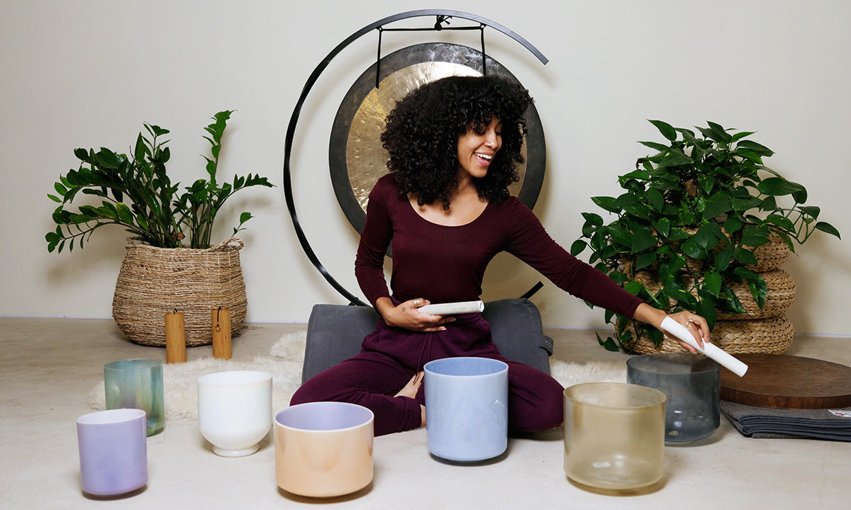 A person sits on a soft rug surrounded by various colored sound bowls and plants, preparing to play singing bowls.