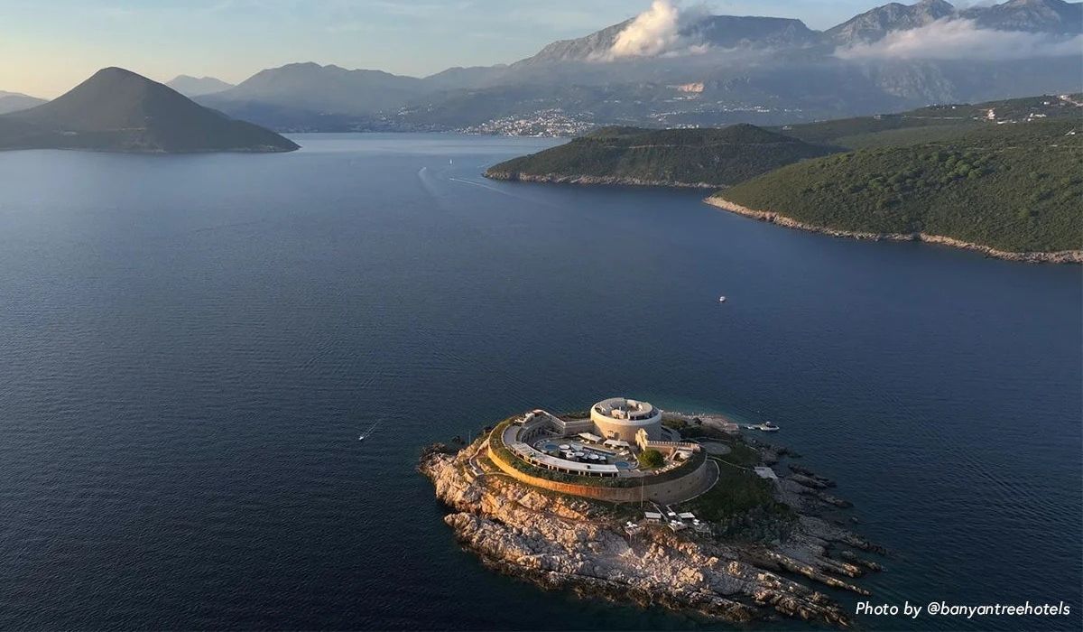 Aerial view of a circular fortress on a rocky islet surrounded by calm blue sea with mountains and scattered clouds in the background.