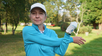 A person in a blue sweater and golf gloves stretches their arm in a grassy park, surrounded by trees.