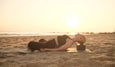 A person practicing yoga on a beach at sunset, lying back on a mat with a block for support, surrounded by soft sand.