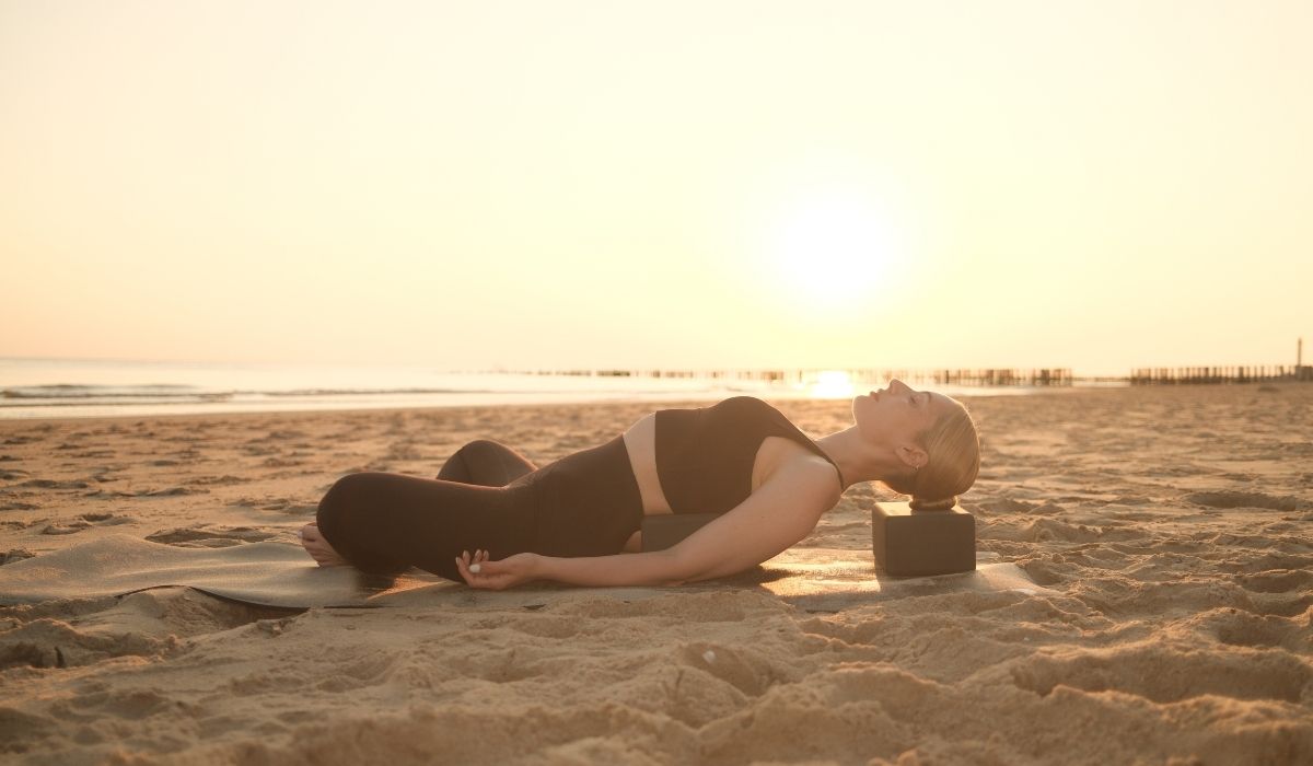 A person practicing yoga on a beach at sunset, lying back on a mat with a block for support, surrounded by soft sand.