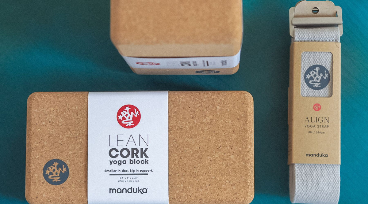 A cork yoga block and a gray yoga strap, both by Manduka, displayed on a blue surface, highlighting eco-friendly fitness accessories.